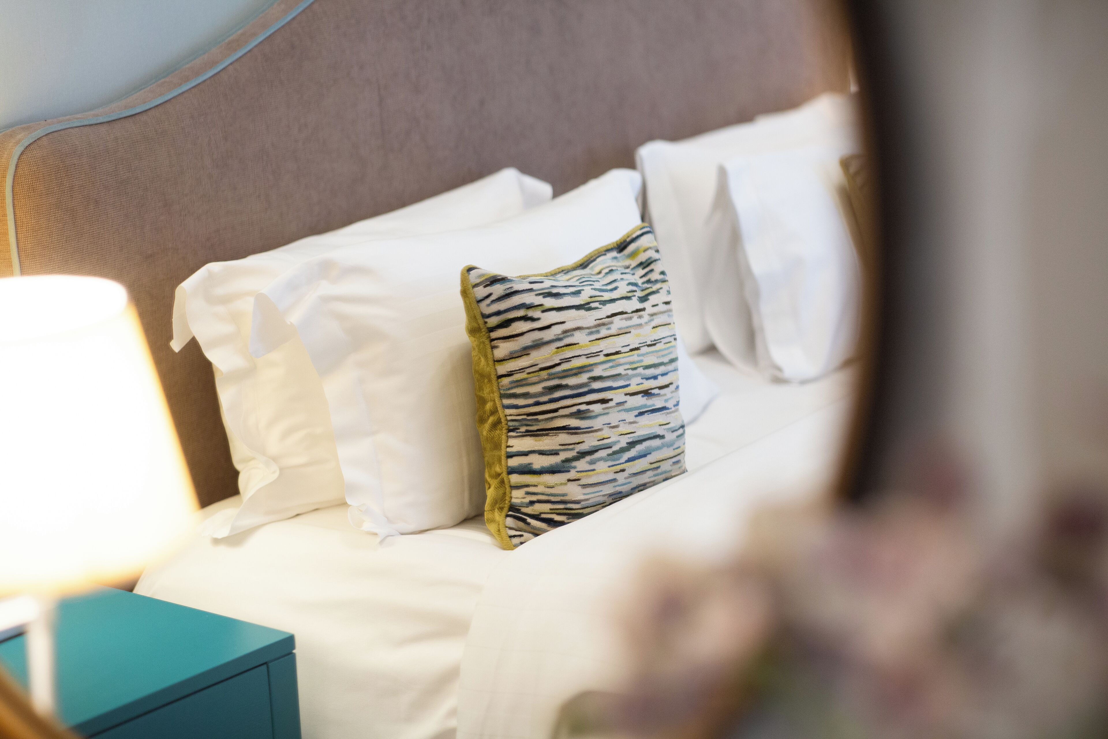 A neatly made bed with white linens and a patterned pillow, a teal bedside table, and a wall with a textured finish.