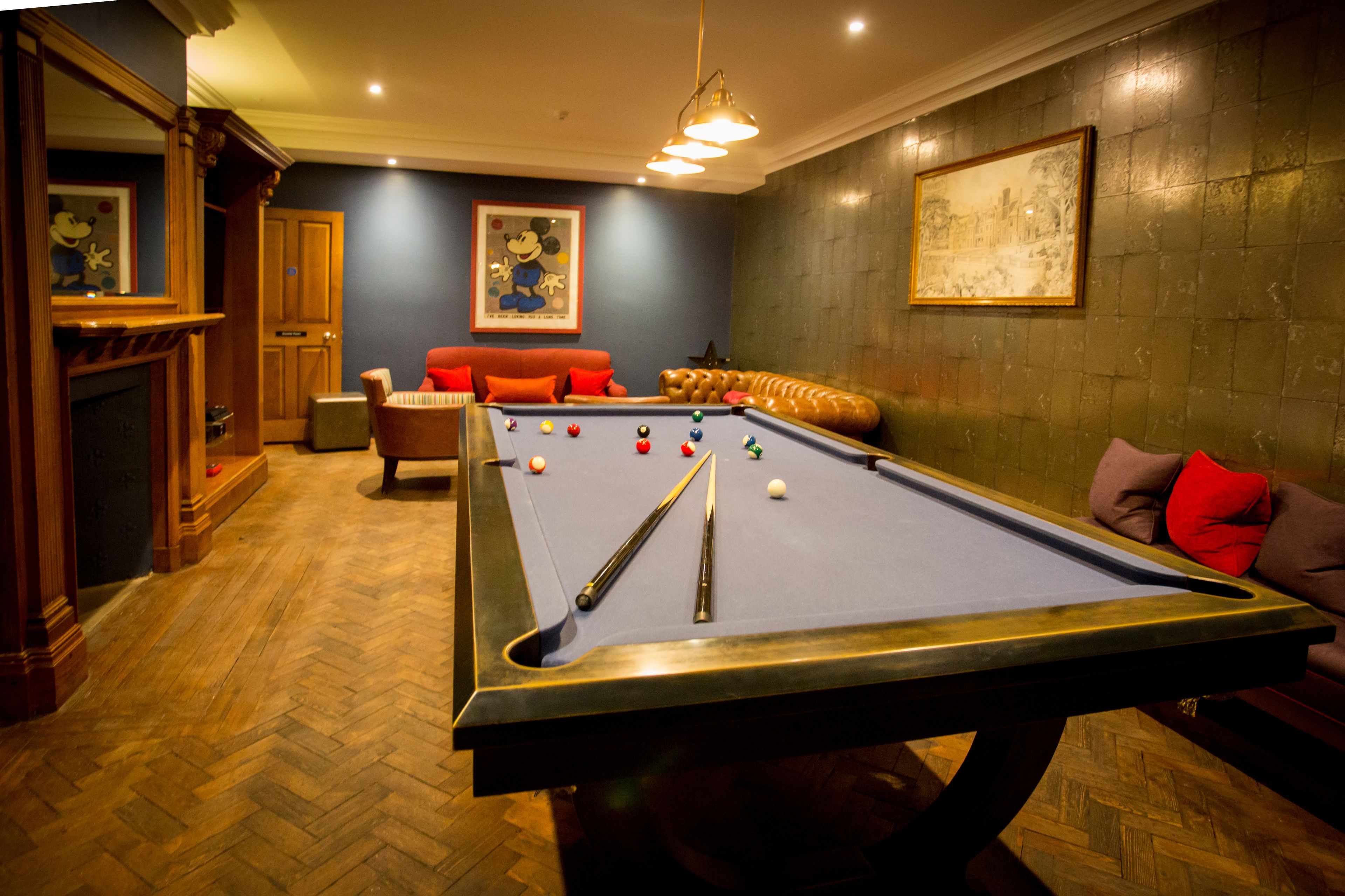 A pool table in a room with a herringbone floor, a fireplace, and a sofa with red cushions.