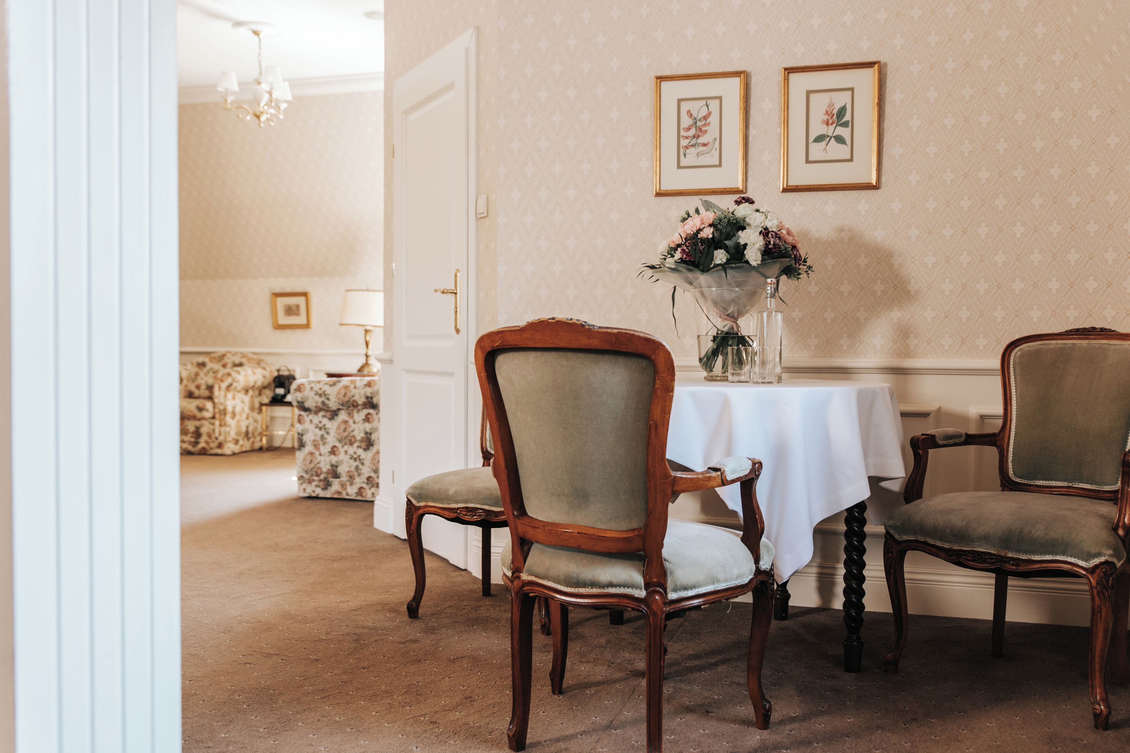 A dining area with a table set for four, surrounded by four wooden chairs with cushioned seats. There is a vase with flowers on the table, and two framed pictures on the wall above.