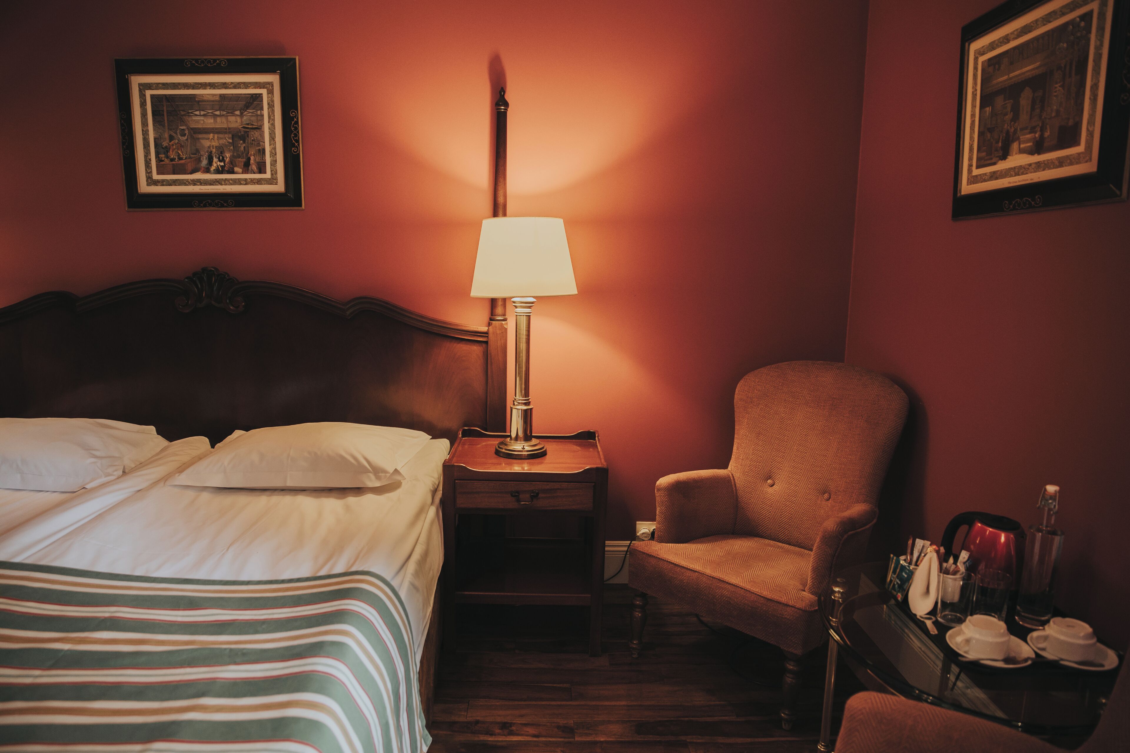 Habitación de hotel con cama, una silla, mesita de noche con lámpara y bandeja con tazas y una botella.