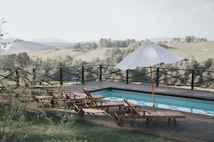Área da piscina com espreguiçadeiras e guarda-sóis de madeira, ladeada por um deck de madeira e vista para colinas ondulantes.