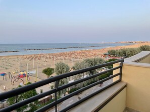 A beach view from a balcony with a railing, a netted volleyball court, and a Ferris wheel in the distance. - Hotel Costazzurra (Rimini)