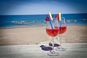 Two glasses of red beverage with fruit garnishes on a wooden table overlooking a beach. - Hotel Costazzurra (Rimini)