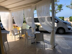 An outdoor seating area with white chairs and tables, a covered section with white curtains, and a view of a parked car and a bus in the background. - Hotel Costazzurra (Rimini)