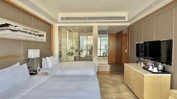 A hotel room with two beds, a TV, a minibar, and a view of the city through a glass door.