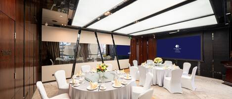 Un salón de conferencias con mesas redondas, sillas blancas y una pantalla grande.