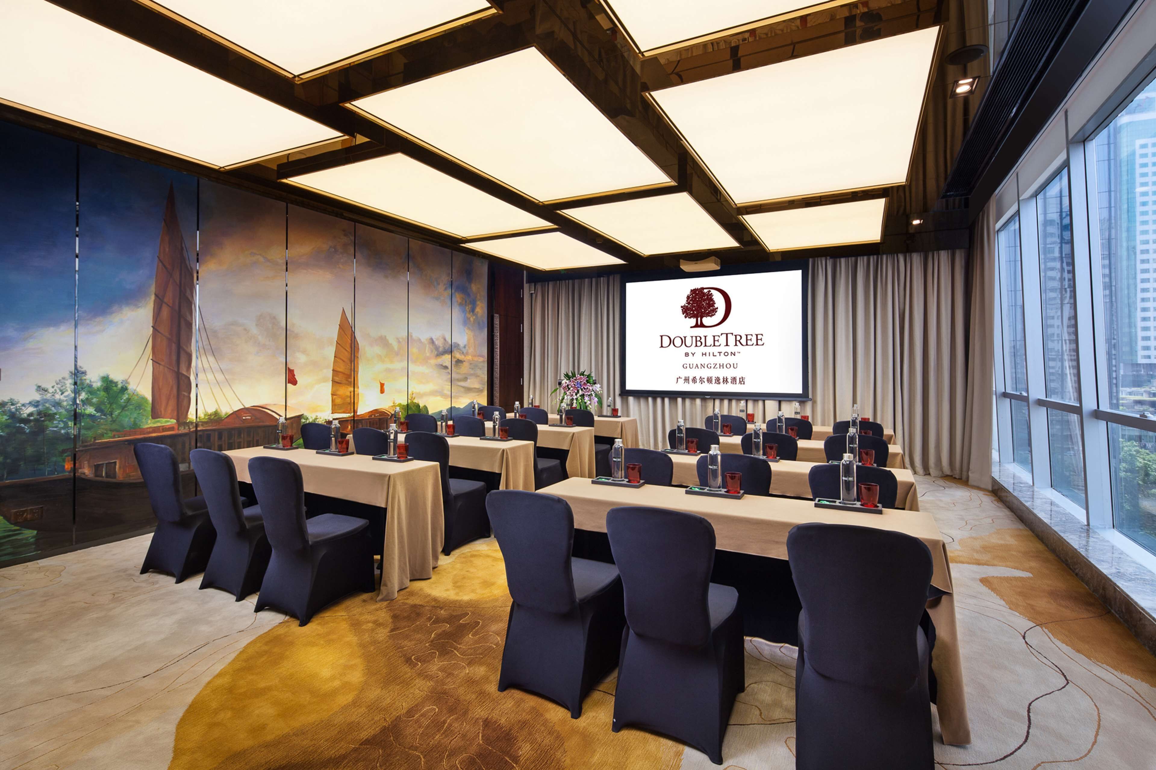 Une salle de conférence dotée d’un grand écran affichant le logo de l’hôtel DoubleTree de Guangzhou, de plusieurs tables avec des chaises, et d’une fresque murale représentant un paysage urbain.