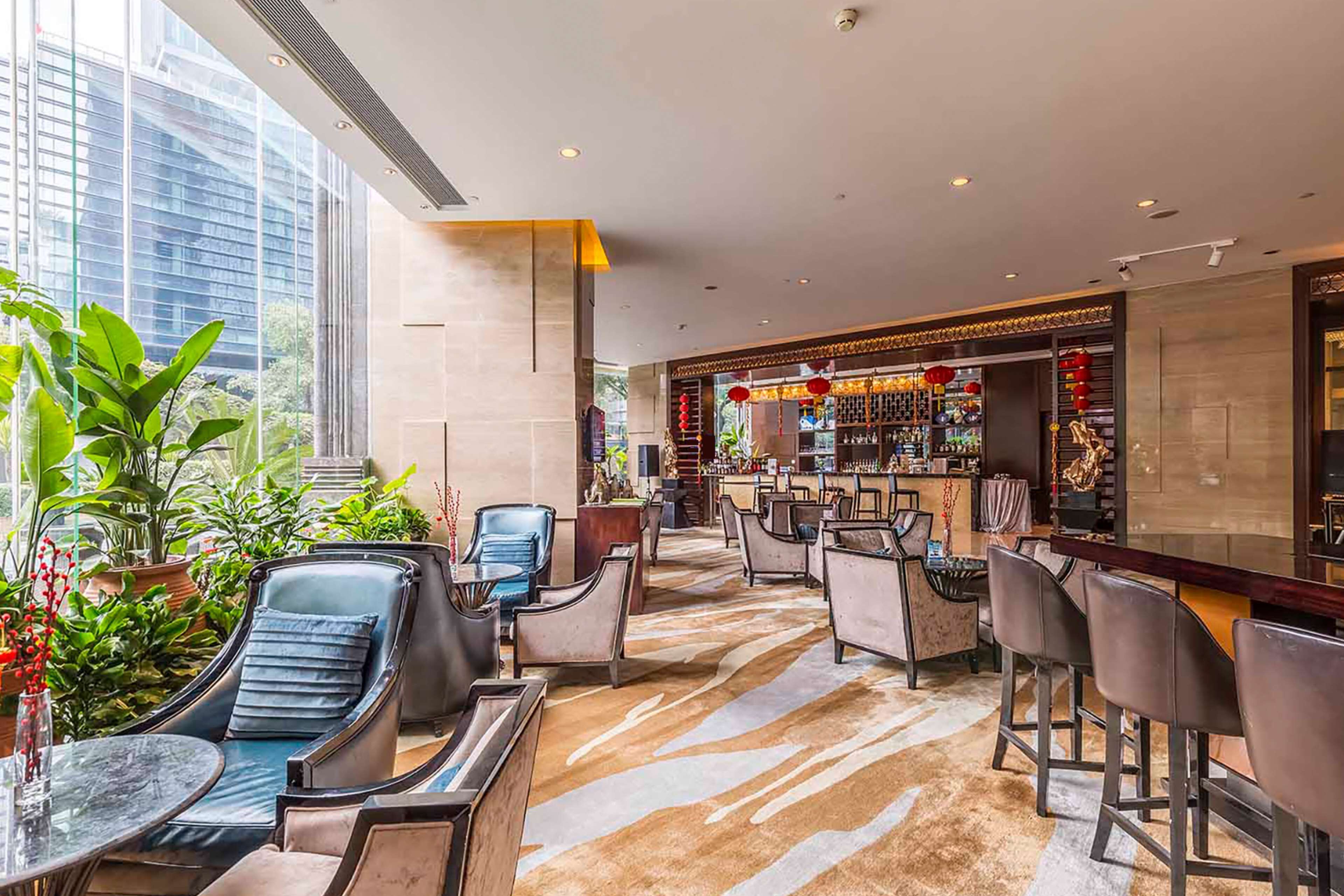 A modern bar area with seating and a bar counter, surrounded by large windows and indoor plants.