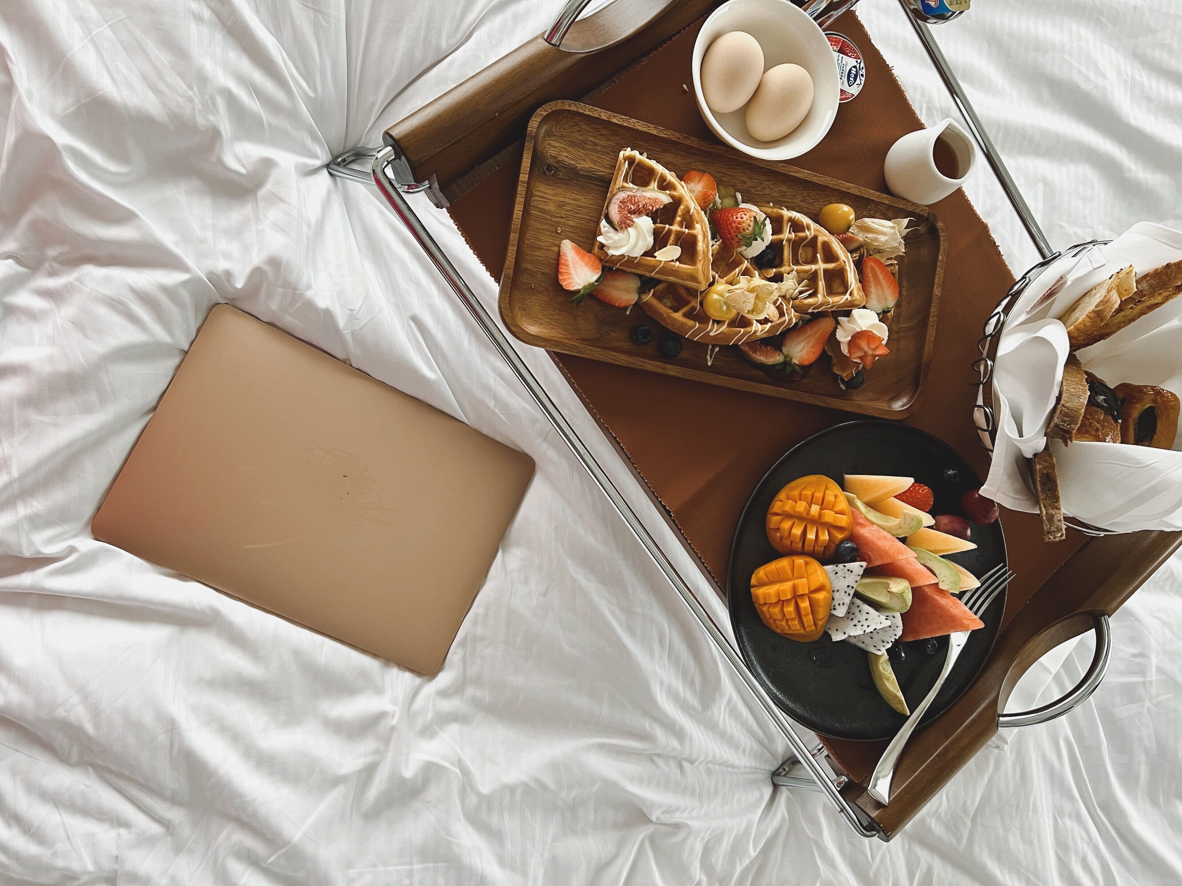 A bed with a laptop, a tray with waffles, fruits, and a cup of coffee.
