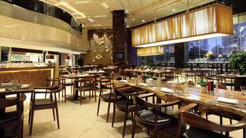 A spacious restaurant with multiple dining tables and chairs, a high ceiling with recessed lighting, and a large window offering a view of the cityscape.