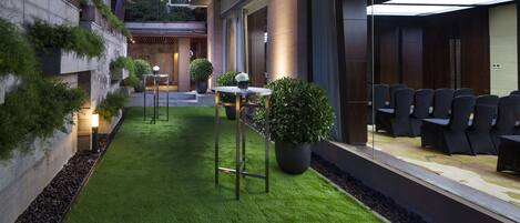 A modern outdoor seating area with artificial grass, potted plants, and a row of chairs.