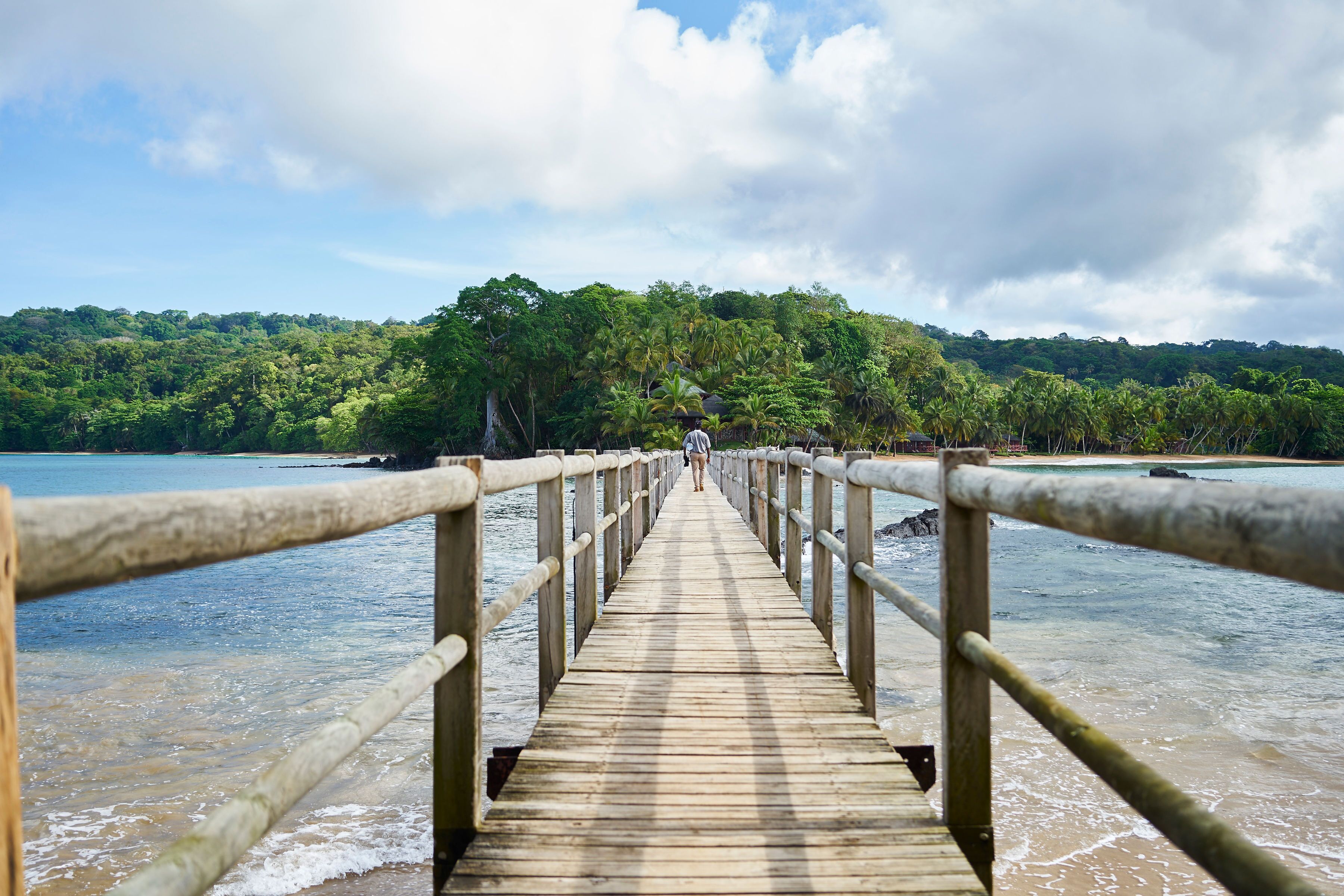 A wooden pier leading to a beach with a forested hill in the background.
