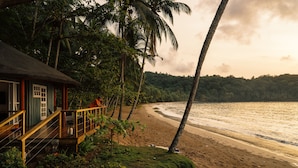 A wooden deck with a railing overlooking a beach, palm trees, and a house with a thatched roof. - Bom Bom – Principe Collection (Príncipe Island)