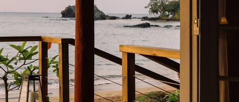 A wooden deck with a railing, a bench, and a view of the ocean with palm trees and a rocky island.