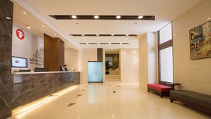 A modern hotel lobby with a reception desk, seating area, and a staircase leading to upper floors.