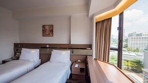 A hotel room with two beds, a nightstand, a bedside table, a painting on the wall, and a window with curtains.