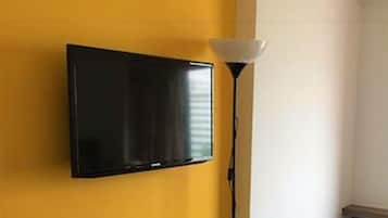 Una habitación con televisión montada en una pared amarilla, una lámpara de pie y una alfombra estampada.