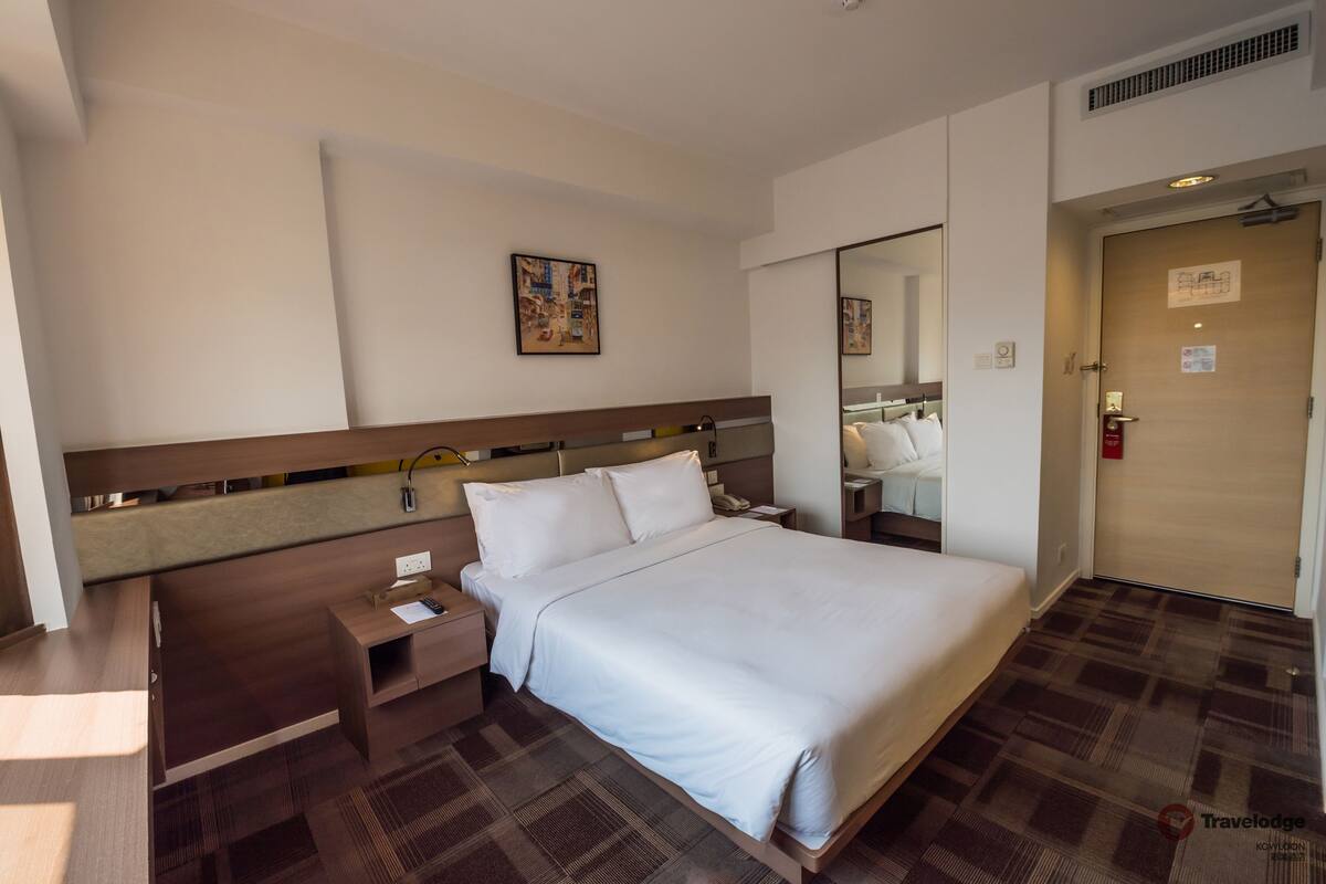 A hotel room with a large bed, a bedside table, a nightstand, a mirror, and a door with a sign.