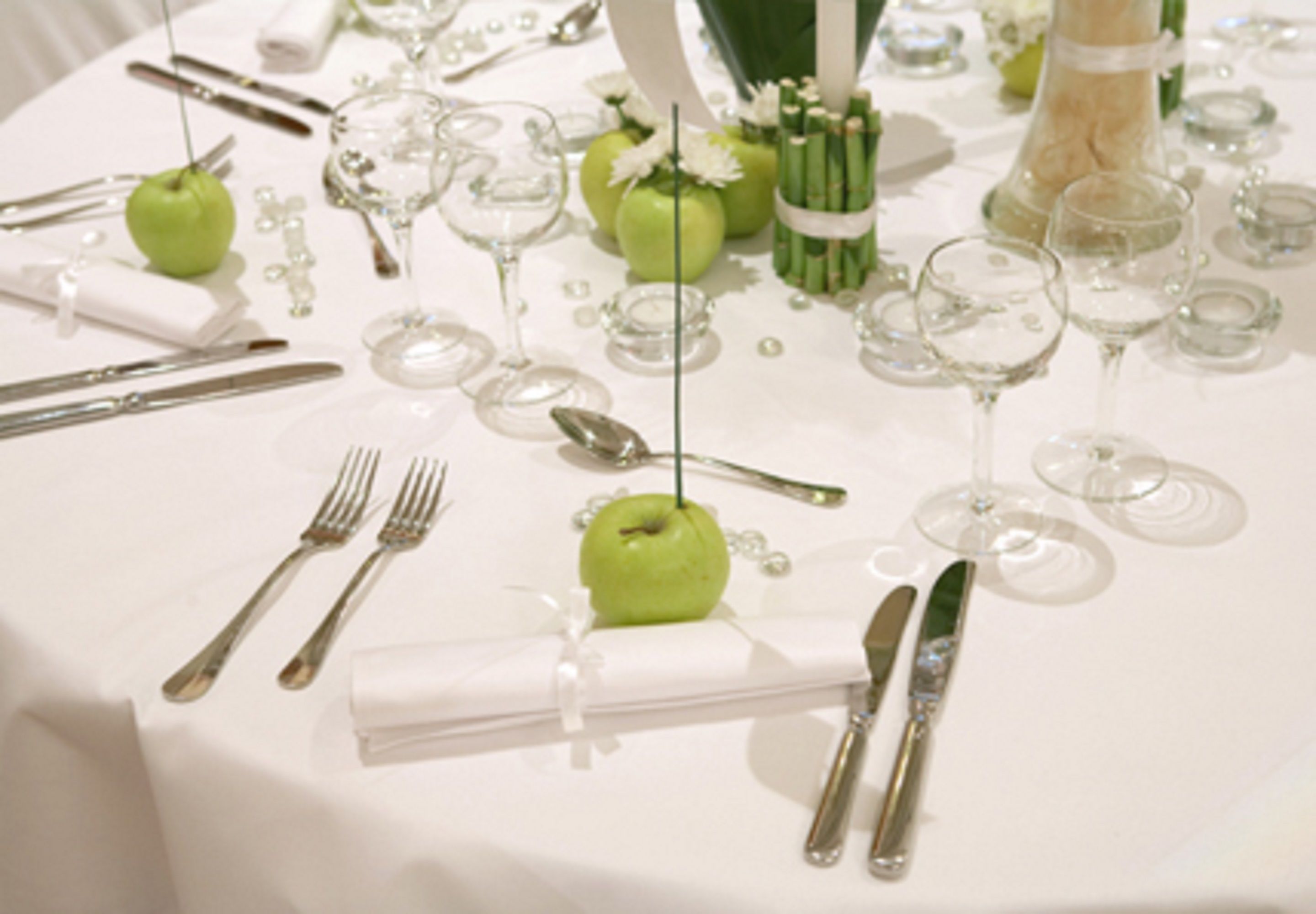 Uma mesa posta com talheres, maçãs verdes, copos de vinho e toalha de mesa branca.