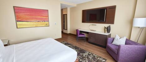 A hotel room with a bed, a desk, a purple sofa, a TV, and a painting on the wall.