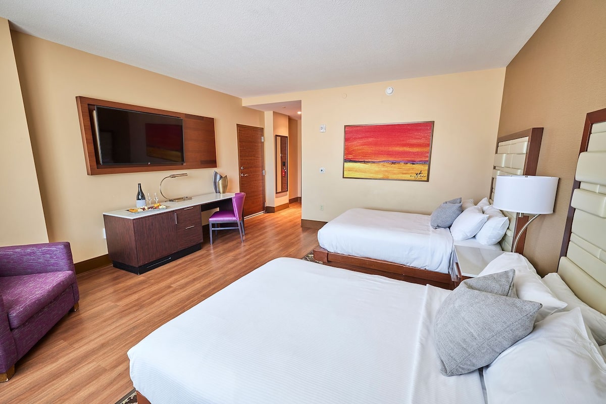 A hotel room with a bed, a desk with a chair, a TV, and a painting on the wall.