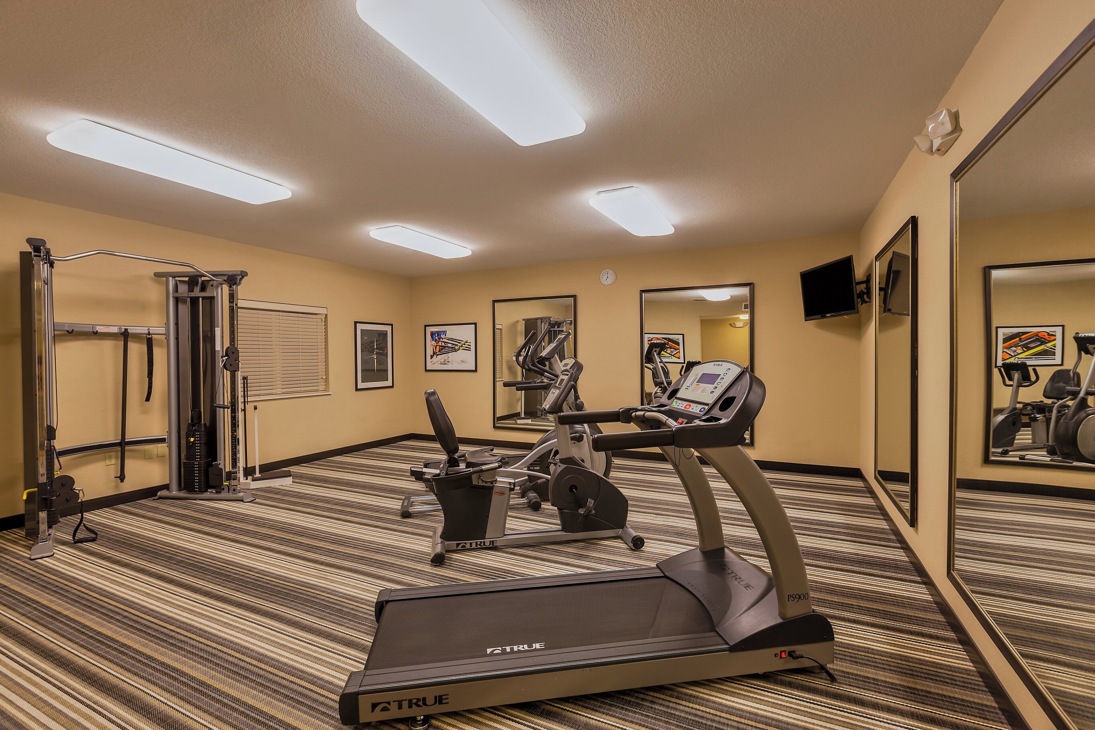 A hotel gym with exercise equipment, including a treadmill and a weight machine, carpeted flooring, and mirrors on the walls.