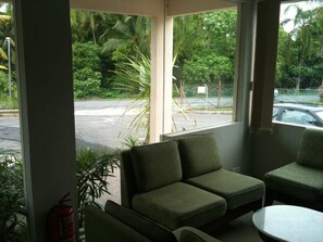 A room with two green cushioned chairs and a small round table, a fire extinguisher, and a view of a parking area with a fence and trees. - Amara Guest House (Langkawi)