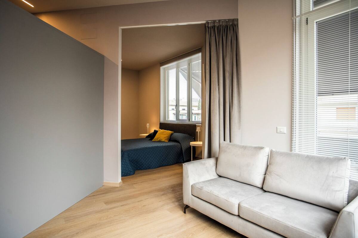 A modern hotel room with a sofa, a bed with a blue quilt, and a window with blinds.