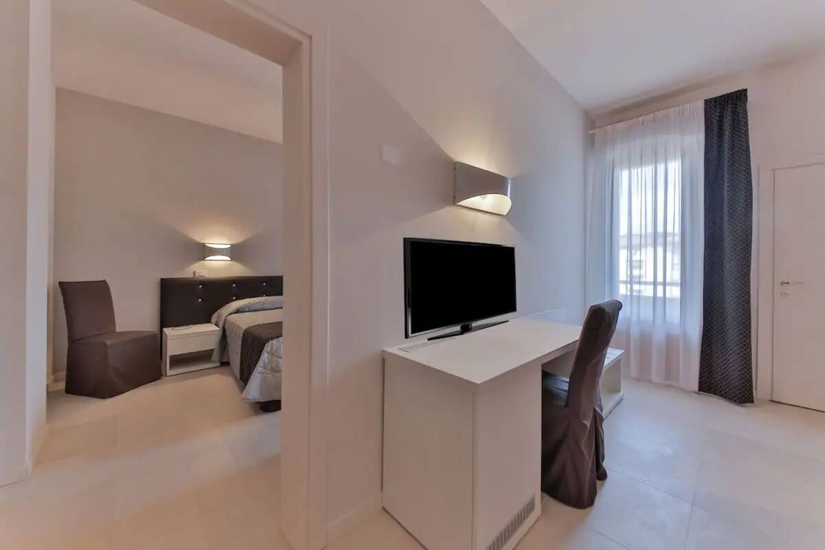A modern hotel room with a bed, bedside table, lamp, TV, desk, chair, and a large window with curtains.