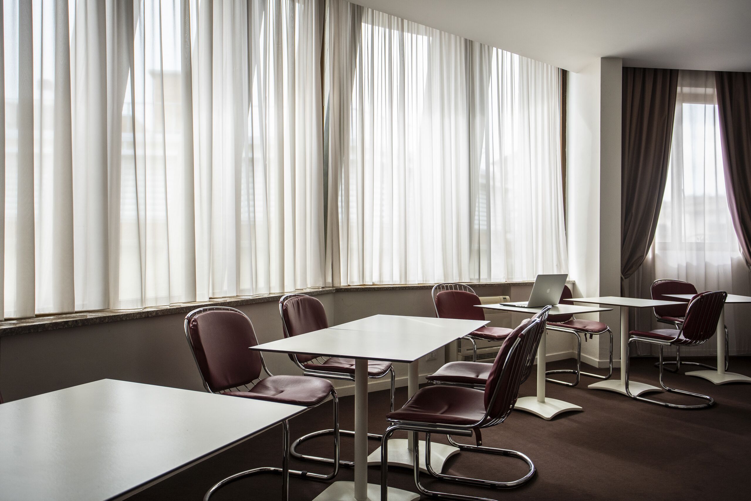 Une salle de conférence avec des tables et des chaises blanches, de grandes fenêtres ornées de rideaux légers et un ordinateur portable posé sur l’une des tables.