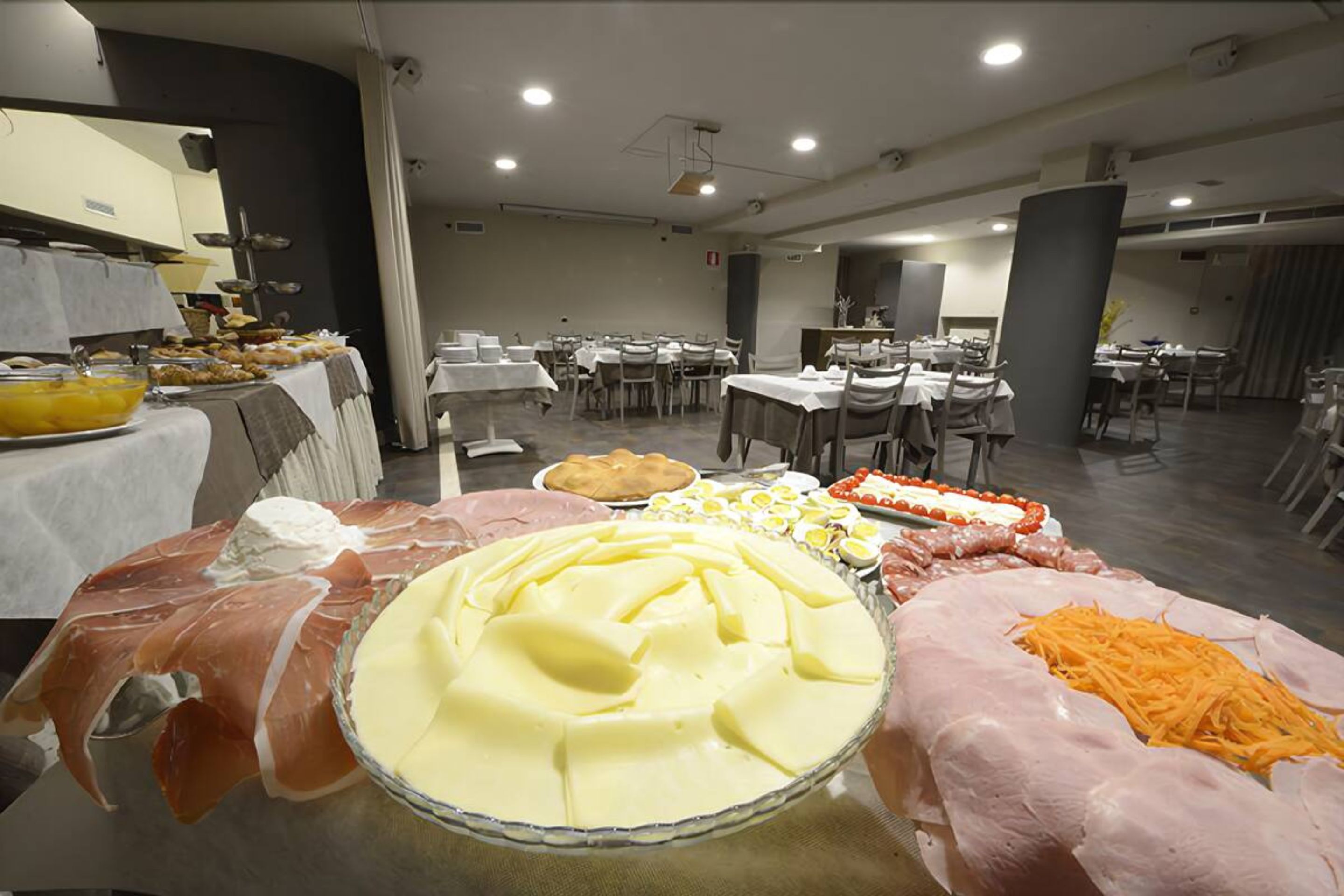 A buffet setup with various food items including cheese, meats, and eggs. Tables are arranged in a spacious room with overhead lighting.