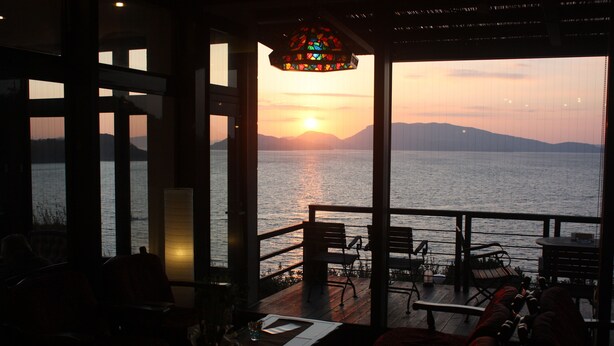 A dining area with a view of the sunset over the water, featuring a wooden deck and outdoor seating.