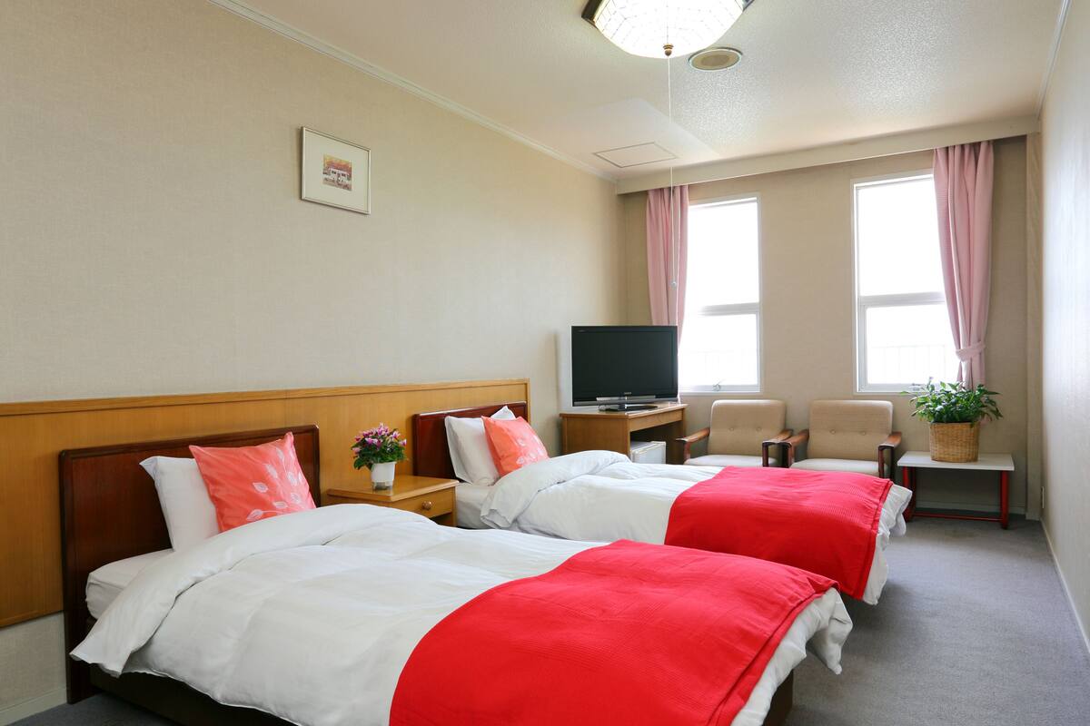 Quarto de hotel com duas camas, televisão, escrivaninha, cadeira e janela com cortinas.