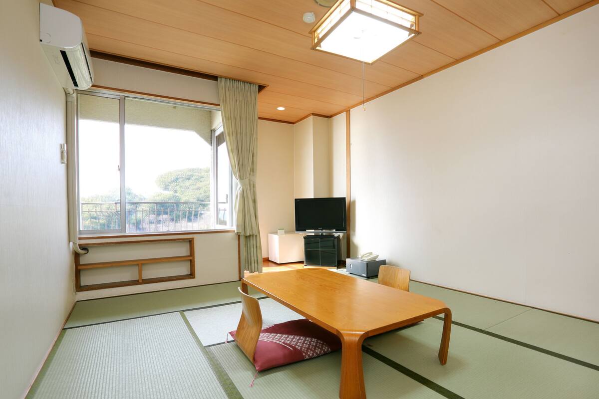 Quarto com piso de esteira de tatame, uma mesa de madeira, uma televisão e uma janela com vista para vegetação.