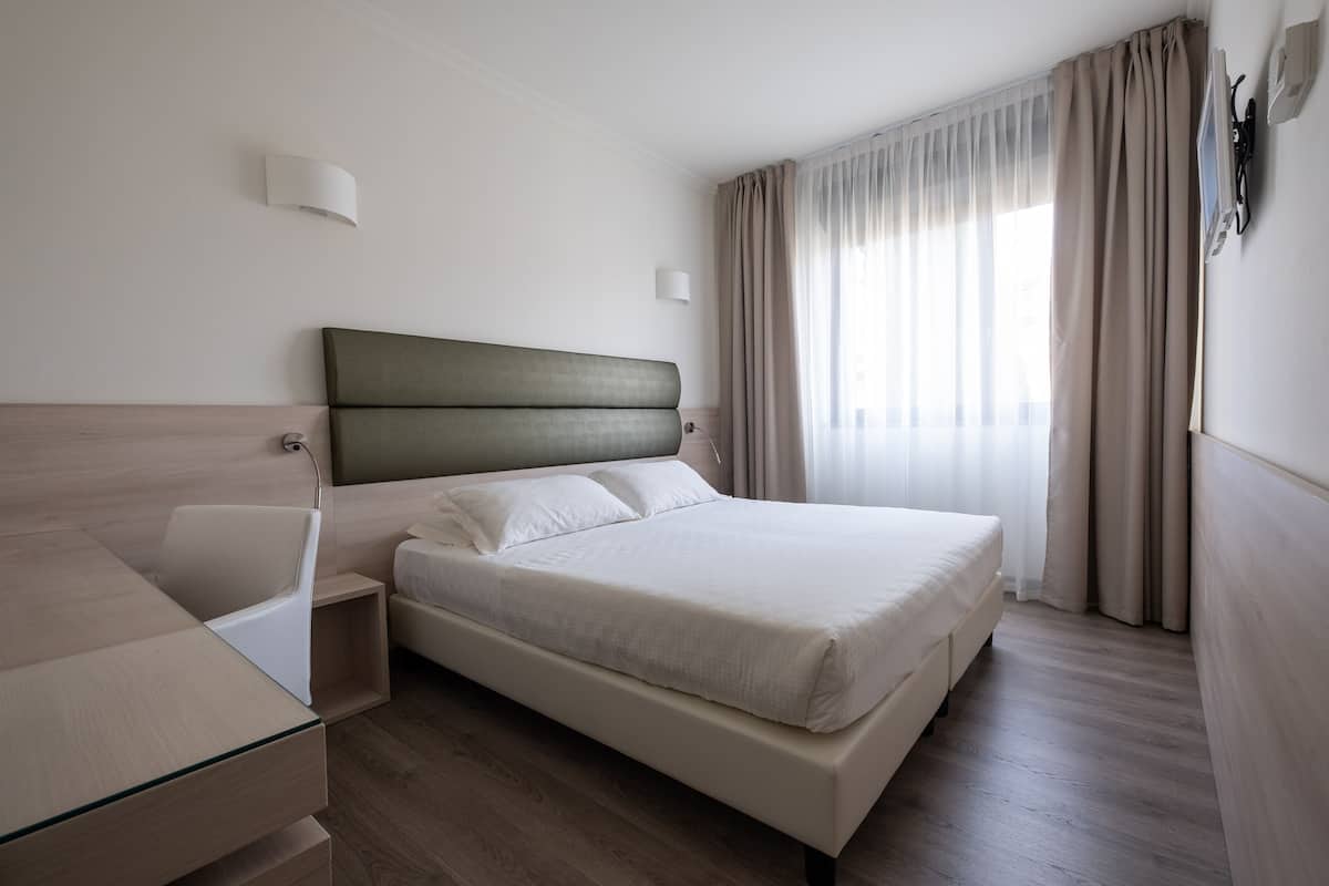 Una moderna camera d'hotel con un letto, un comodino, una lampada e vista sull'esterno attraverso tende leggere.