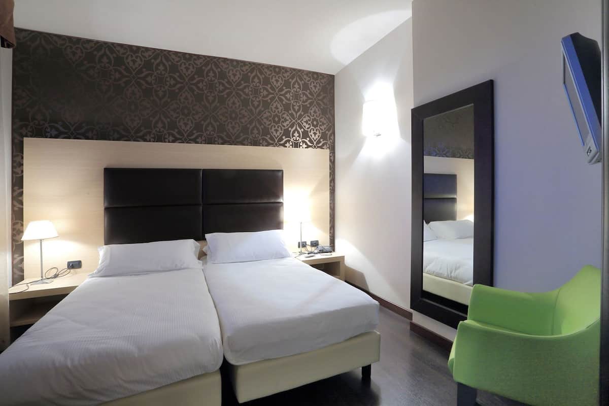 Una camera d'albergo con due letti, uno specchio grande, una sedia verde e una parete decorata.