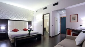 Quarto de hotel moderno com uma cama grande, mesas de cabeceira, um banco, um sofá e uma escrivaninha com cadeira.