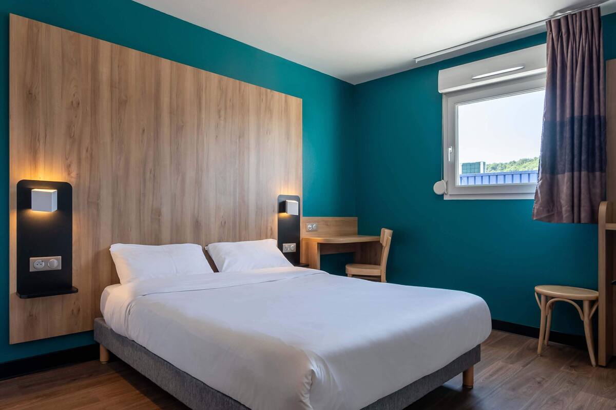 Una habitación de hotel moderna con una cama grande, cabecera de madera, paredes color turquesa y un pequeño escritorio con una silla.