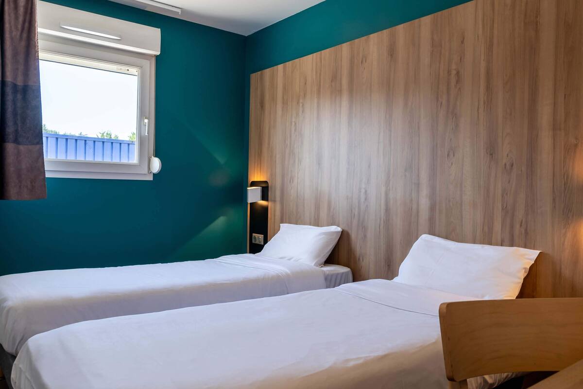 Una habitación con dos camas individuales, una ventana con cortinas, paredes color verde azulado y un muro decorativo de madera.