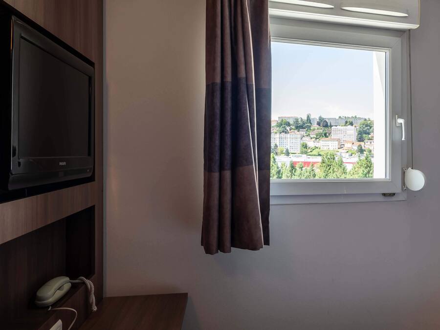 Una habitación con televisor en un estante de madera, teléfono y ventana con cortinas.