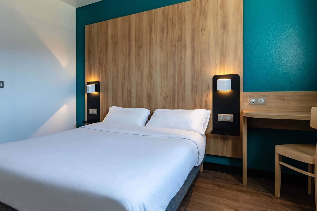 Habitación de hotel con una cama grande, dos mesitas de noche, cabecero de madera y un escritorio con una silla.