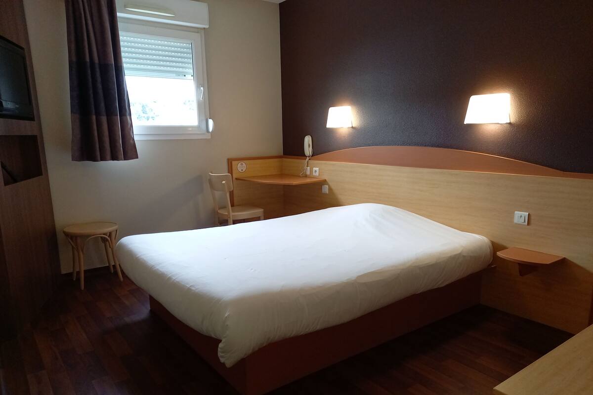 Habitación de hotel con una cama individual, una mesita de noche de madera, una pequeña mesa, una silla y una ventana con persianas.