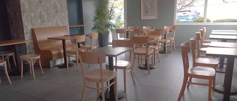 A restaurant with wooden tables and chairs, a large potted plant, and a view of parked cars through the window.