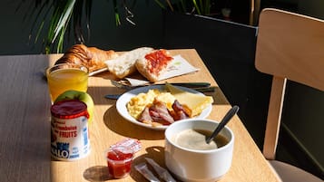 Una mesa de desayuno con croissant, tostadas, huevos revueltos, tocino, un vaso de zumo de naranja, un bol de sopa y una lata de yogur.
