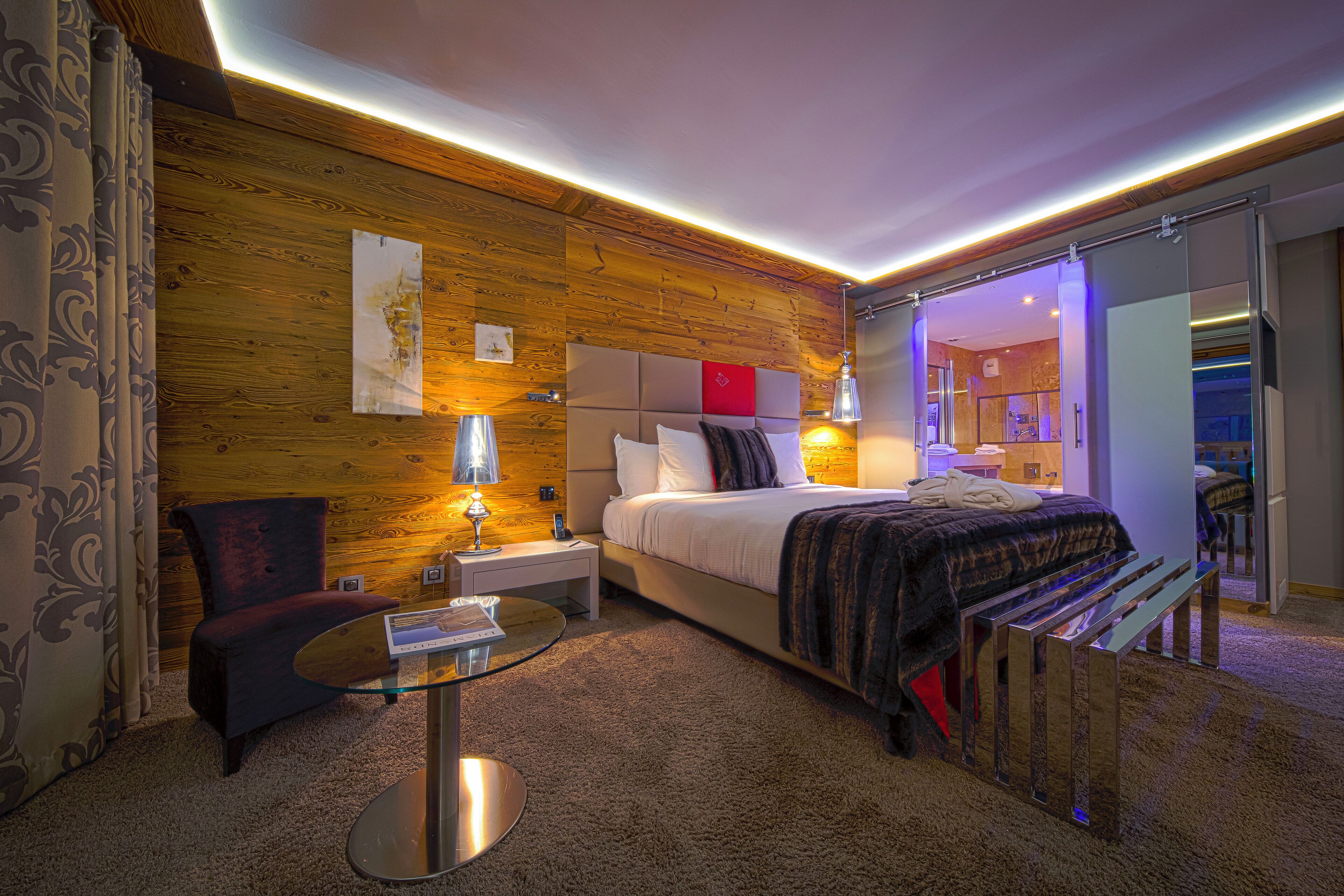 Una habitación de hotel moderna con pared de madera, una cama grande con cabecera, una mesita de noche con lámpara, una pequeña mesa redonda y un banco.
