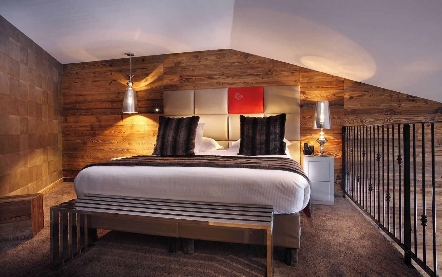 A modern bedroom with a sloped ceiling, wooden panel walls, a large bed with a headboard, bedside lamps, and a nightstand.