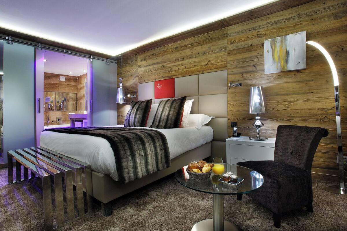 Una habitación de hotel moderna con cama, mesita de noche, silla y una mesa de vidrio con productos para el desayuno.