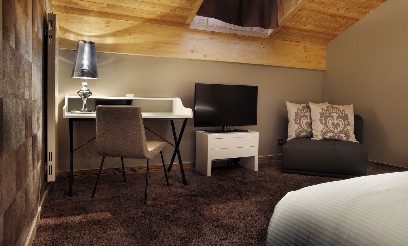 A hotel room with a wooden ceiling, a desk with a lamp, a television, a chair, a bed, and a sofa.