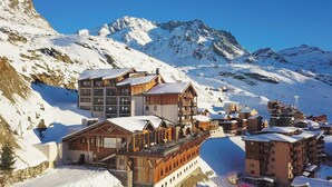 Estância de esqui no alto de uma montanha coberta de neve, com edifícios de madeira, um restaurante e um teleférico ao fundo.