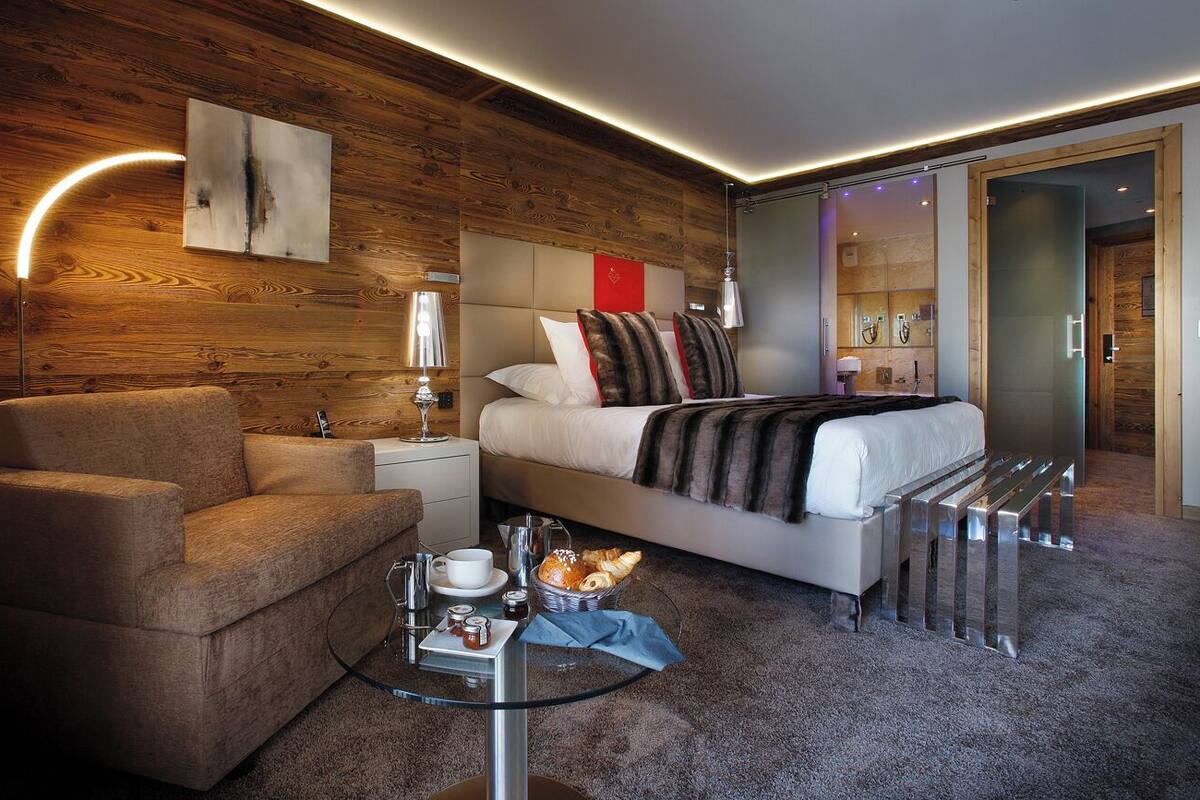 Una habitación de hotel moderna con una pared de madera, una cama con cabecera, una zona de estar con una silla y una mesa de vidrio, y un baño visible a través de una puerta de cristal.